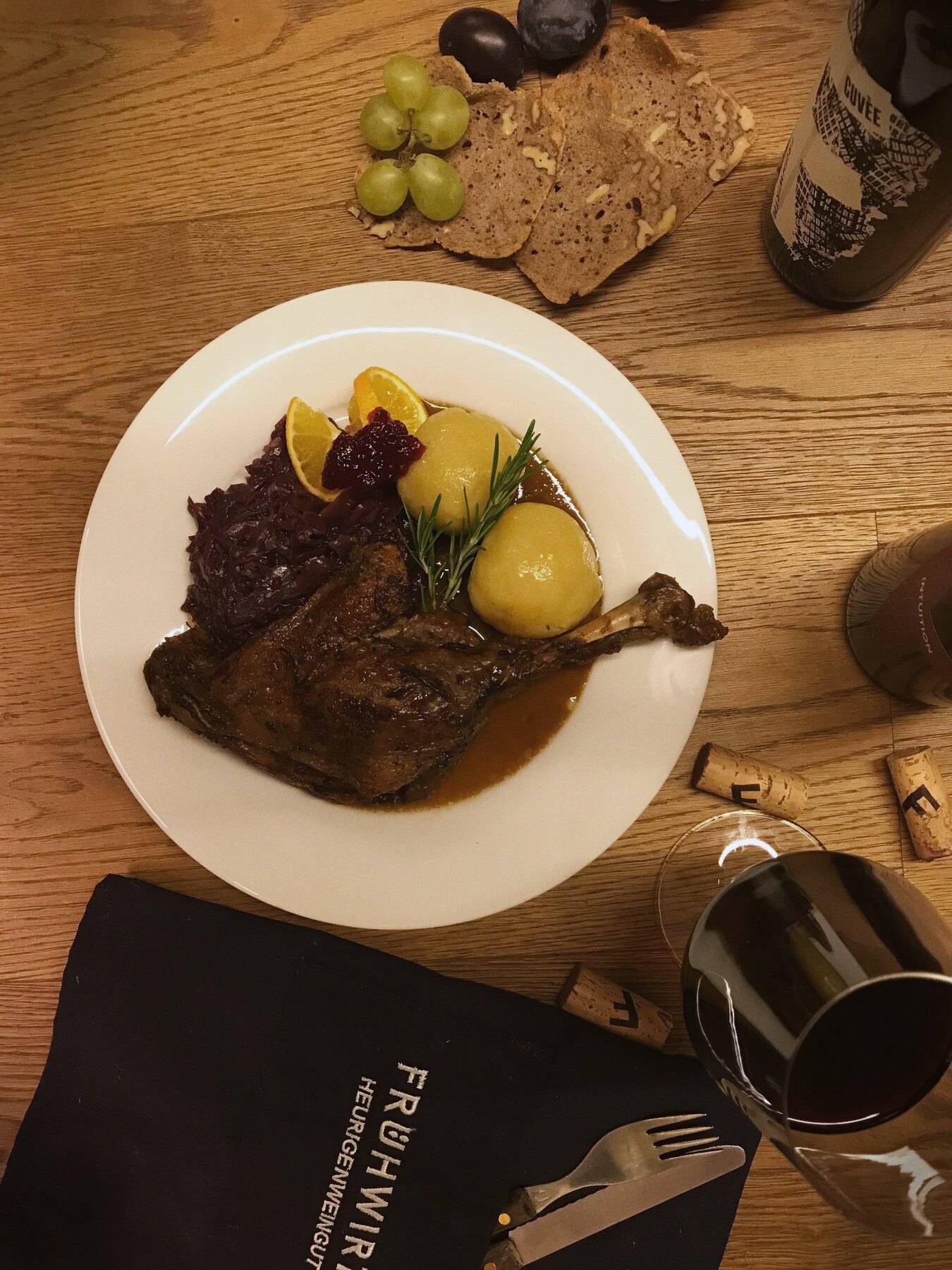 Gänsekeule mit Erdäpfelknödel, Rotkraut, Orange und Preiselbeeren von oben, daneben ein Glas Rotwein
