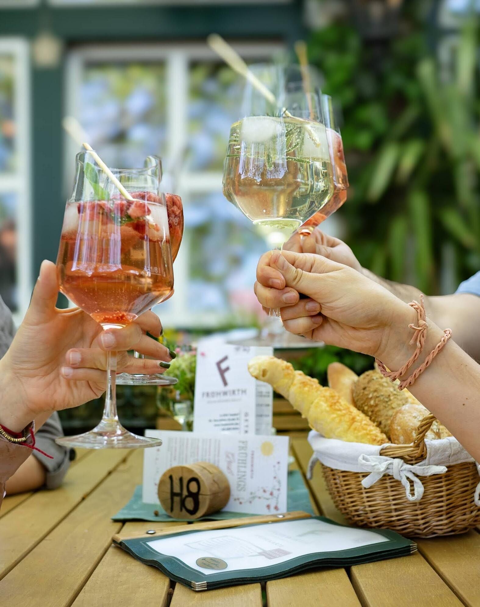 Zwei Hände stoßen im Gastgarten mit einem Rosé-Spritzer und einem Glas Weißwein an, darunter ein Brotkorb am Holztisch