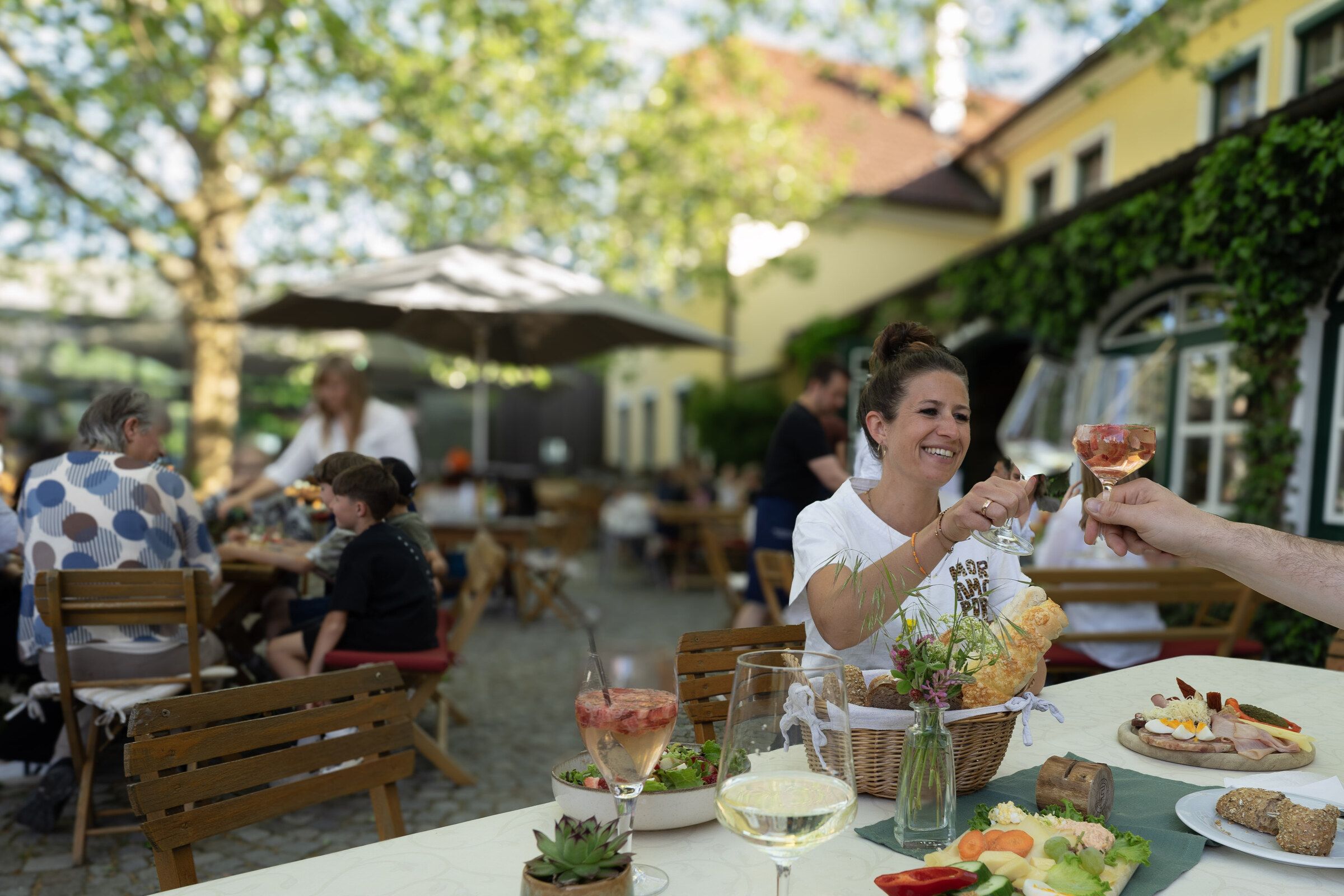 Voller Gastgarten im Mai 2026: eine Frau stößt über den gedeckten Tisch hinweg an