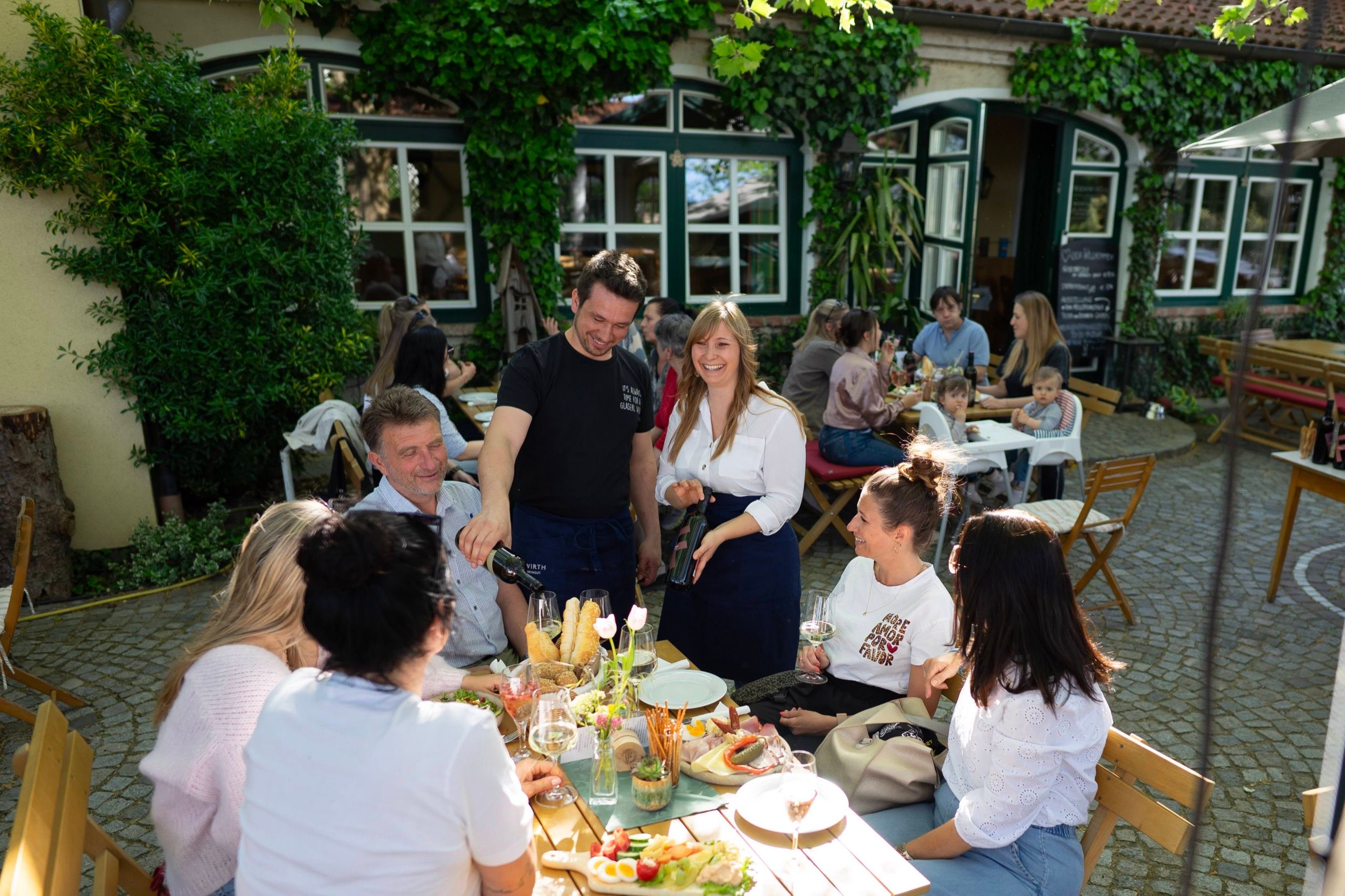 Gastgarten des Heurigen: Service schenkt Wein aus, Gäste an einem langen Tisch