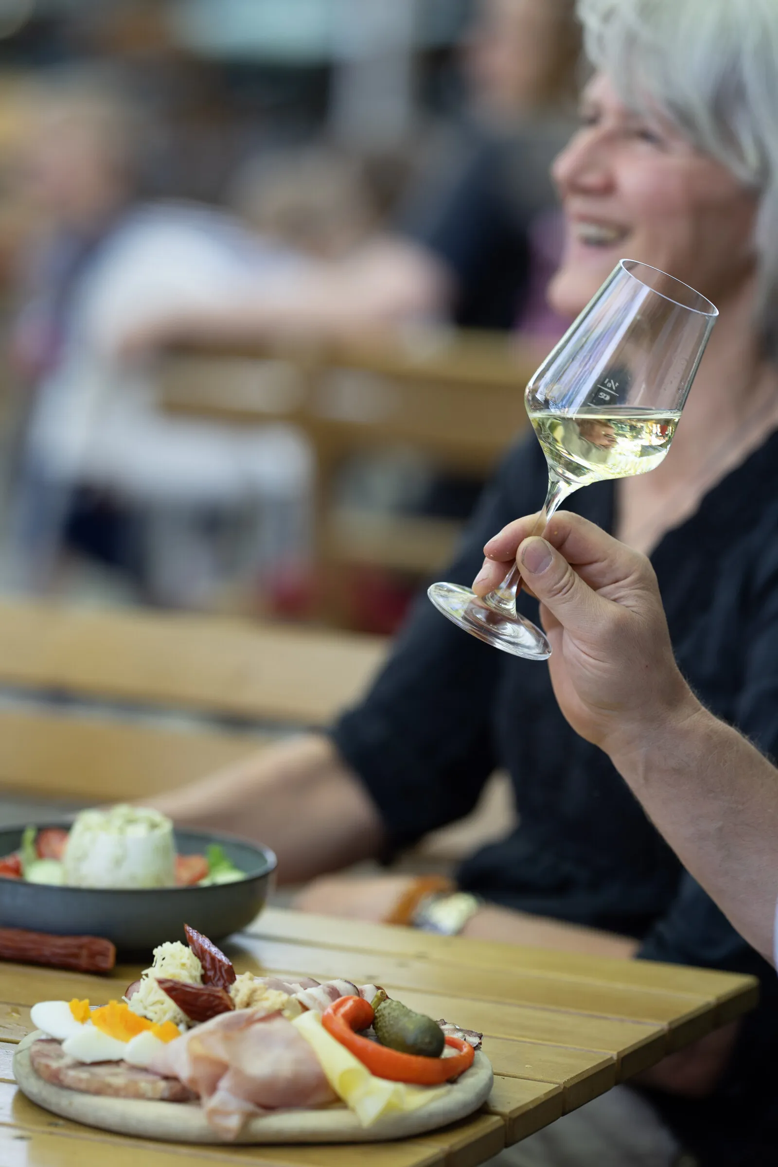A hand raises a glass of white wine, on the wooden table in front a platter with ham, egg and cheese