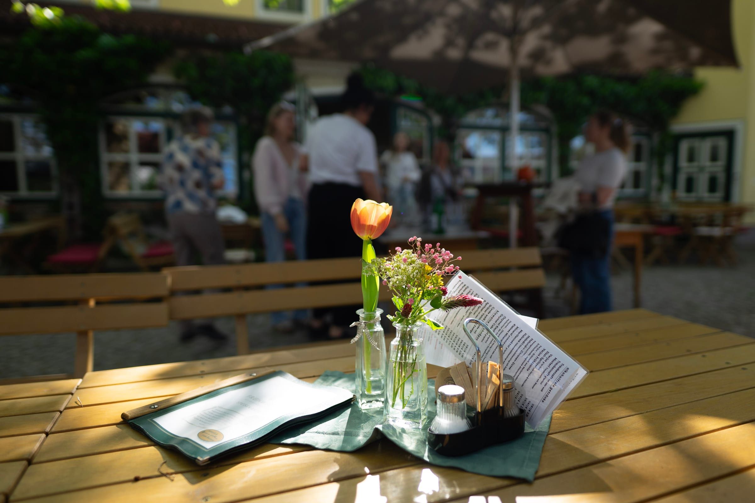 Holztisch mit Tulpe und Speisekarte im Gastgarten, dahinter Gäste unter den Schirmen