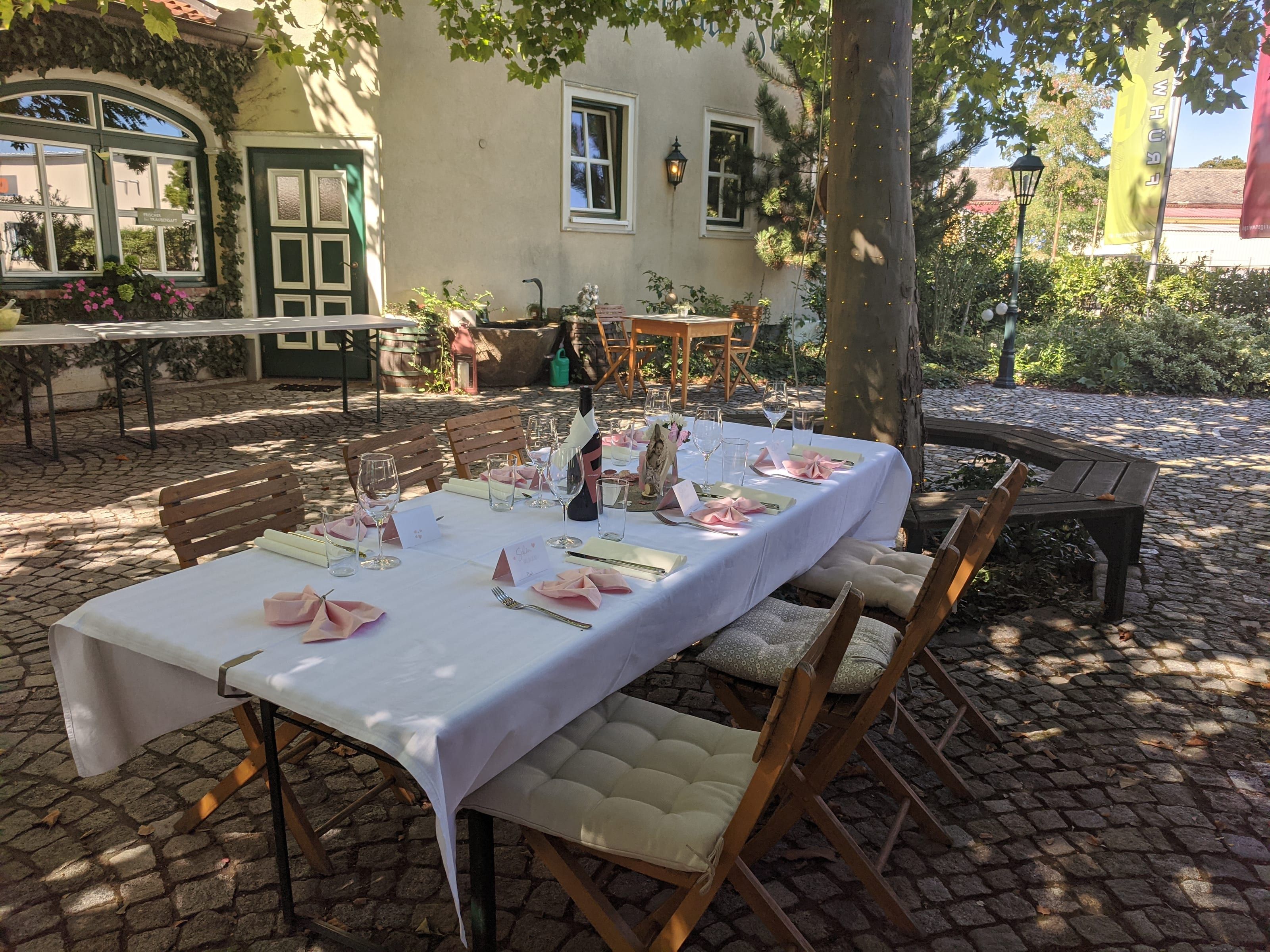 A long festive table laid in white under the tree in the courtyard, the house with green door and window frames behind