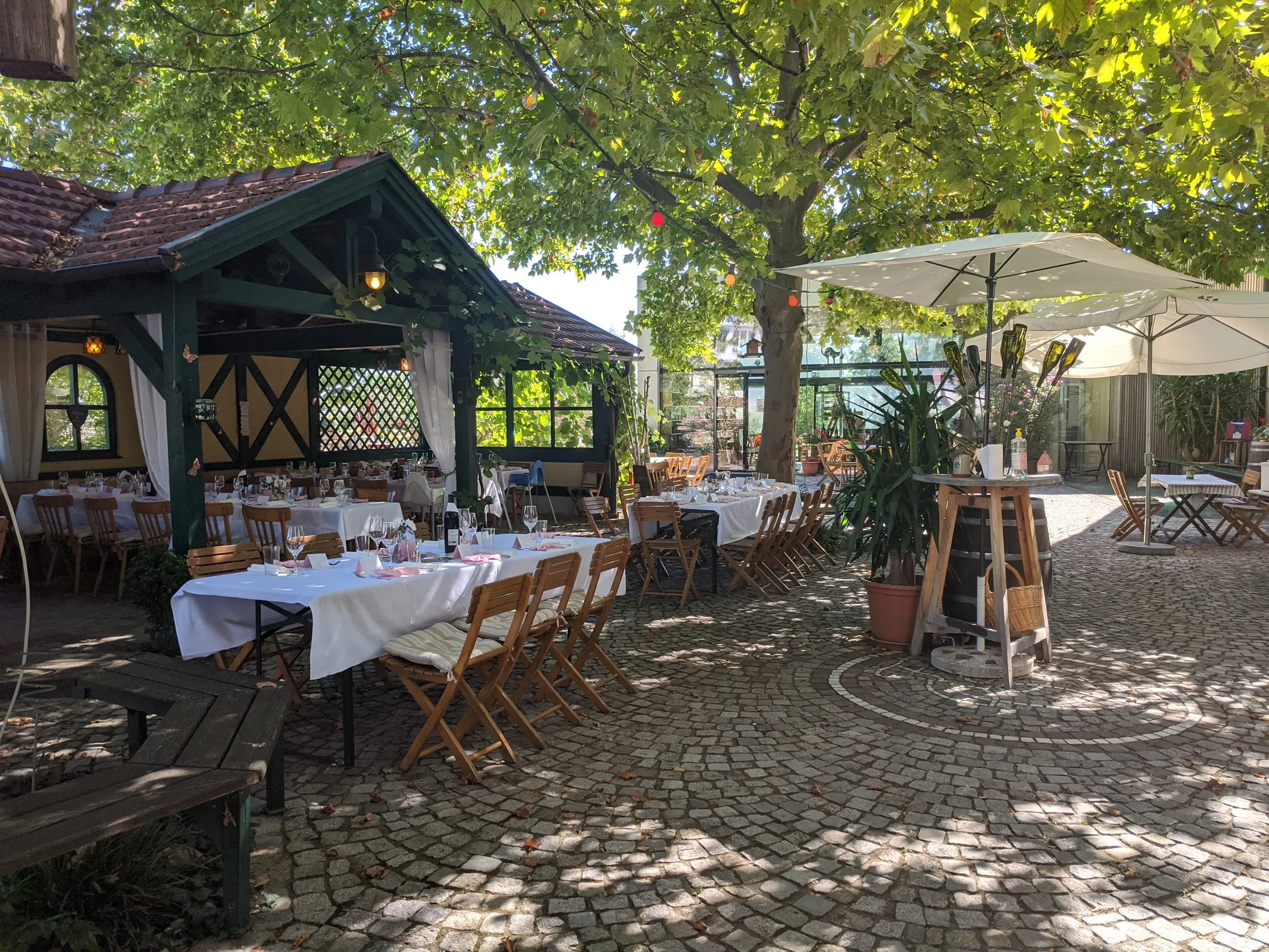 Gastgarten unter alten Platanen: links das offene Salettl mit gedeckten Tischen, rechts ein Sonnenschirm, dazwischen Kopfsteinpflaster