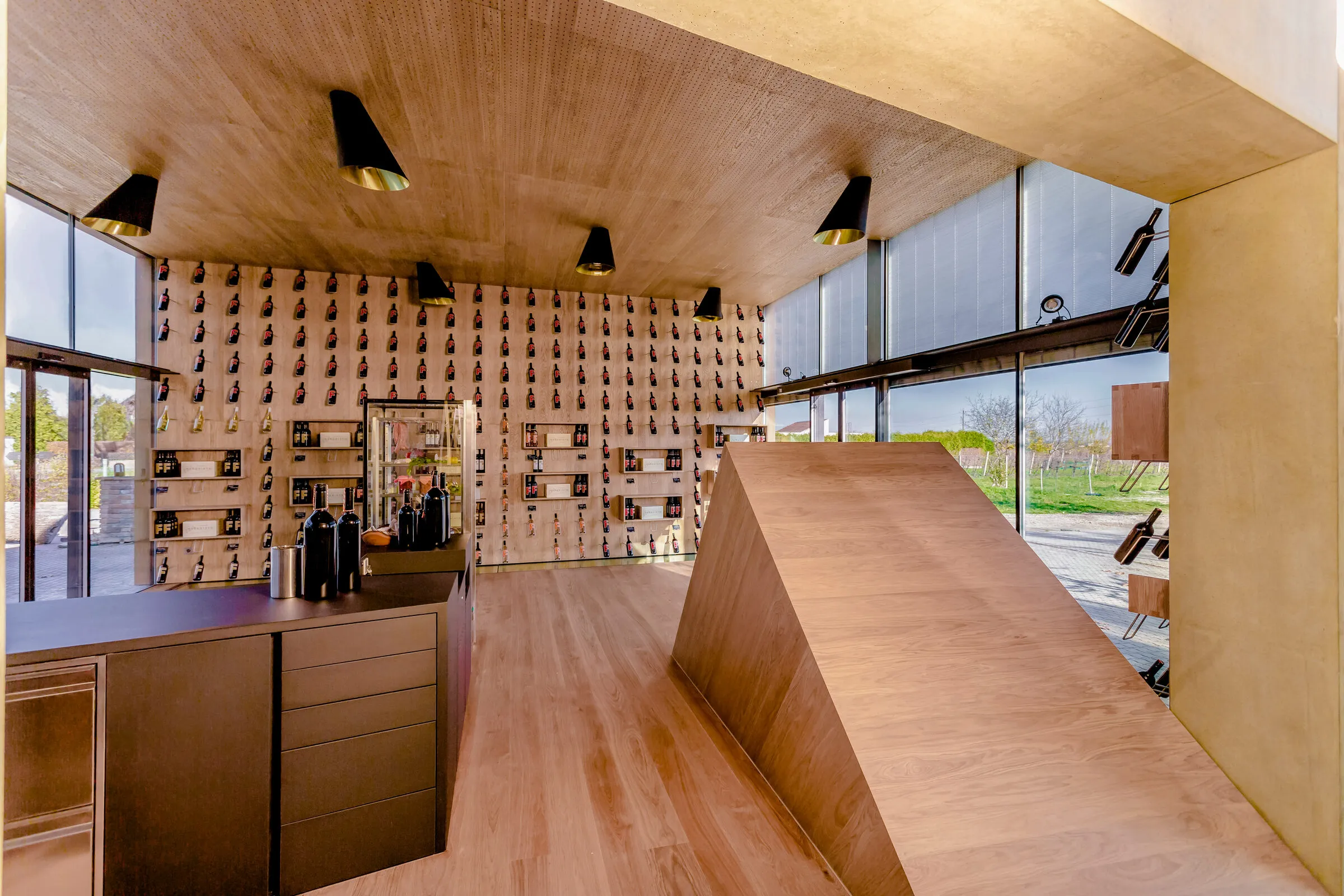 Die Vinothek von der Theke aus: helle Holzrampe, Wand mit gesteckten Flaschen, große Glasfront ins Grüne