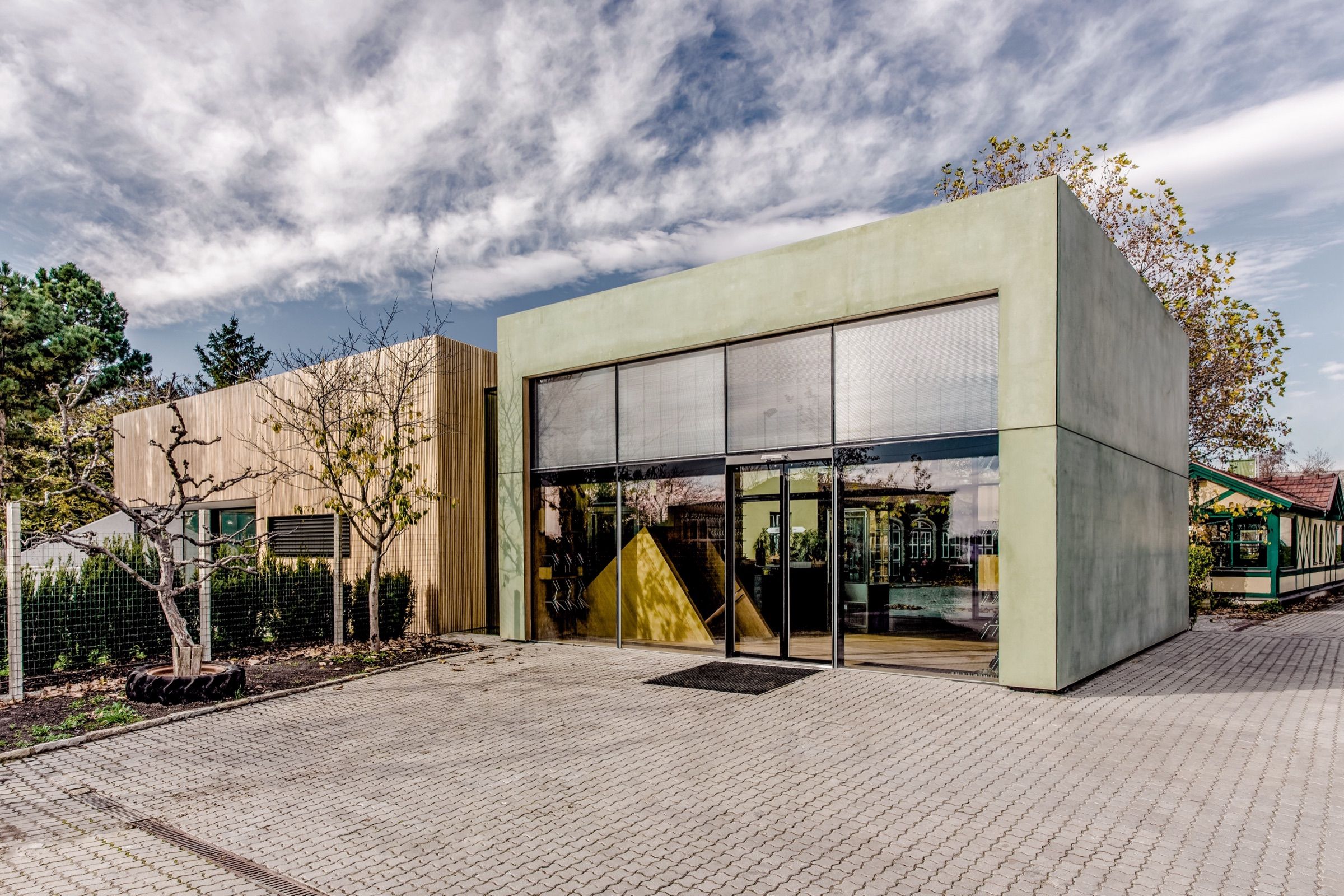 Die Vinothek von außen bei Tag: ein grüner Kubus mit großer Glasfront, links der Holzbau, davor der gepflasterte Hof
