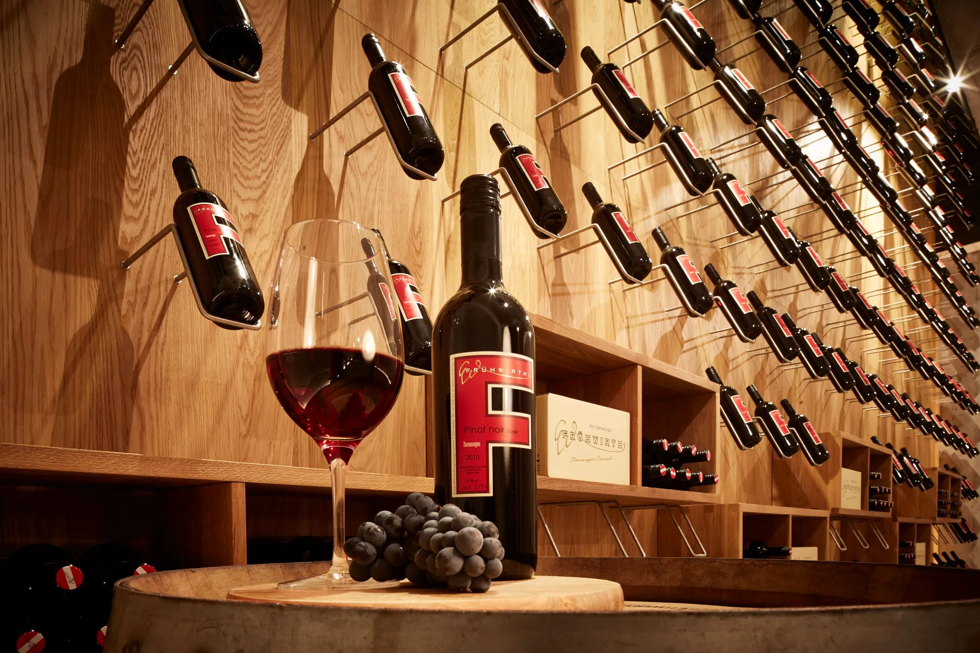Ein Glas Rotwein und eine Flasche vor der Wand mit schräg gesteckten Flaschen