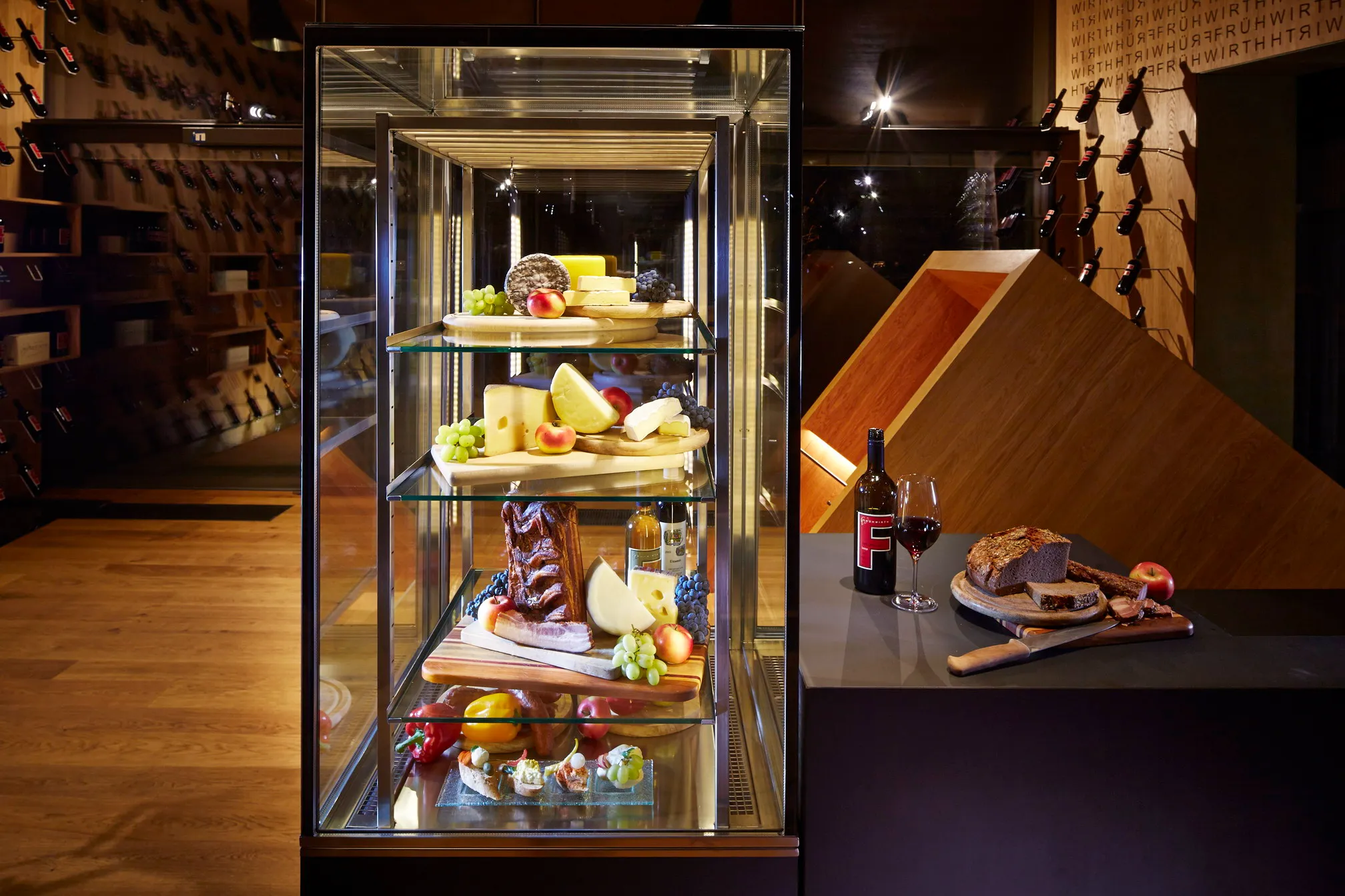 Gläserne Vitrine mit Käse und Aufstrichen, daneben ein Glas Rotwein und Brot