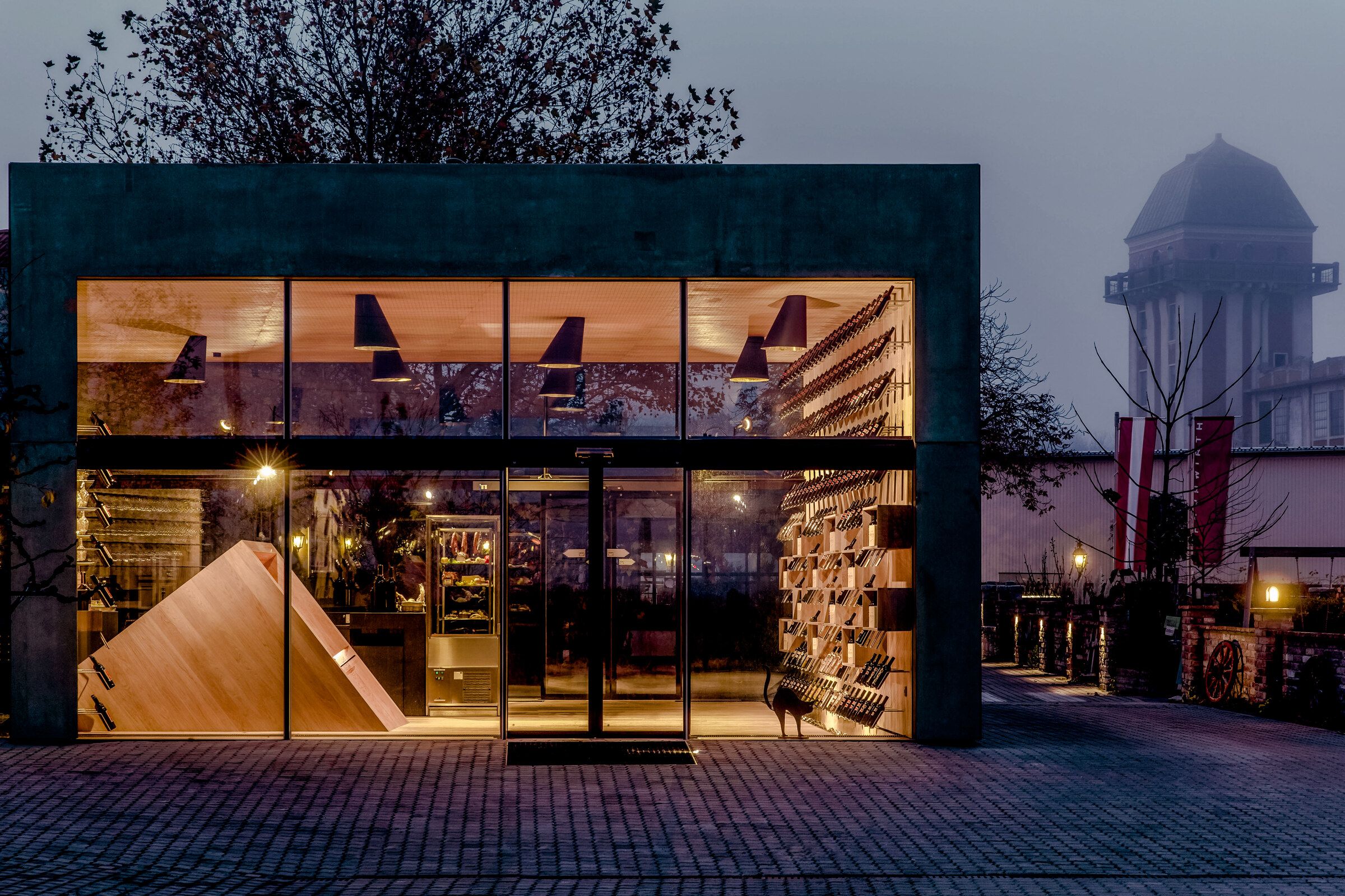 Die Vinothek in der Dämmerung: ein Haus mit großer Glasfront und Holzregalen innen, dahinter der Teesdorfer Wasserturm